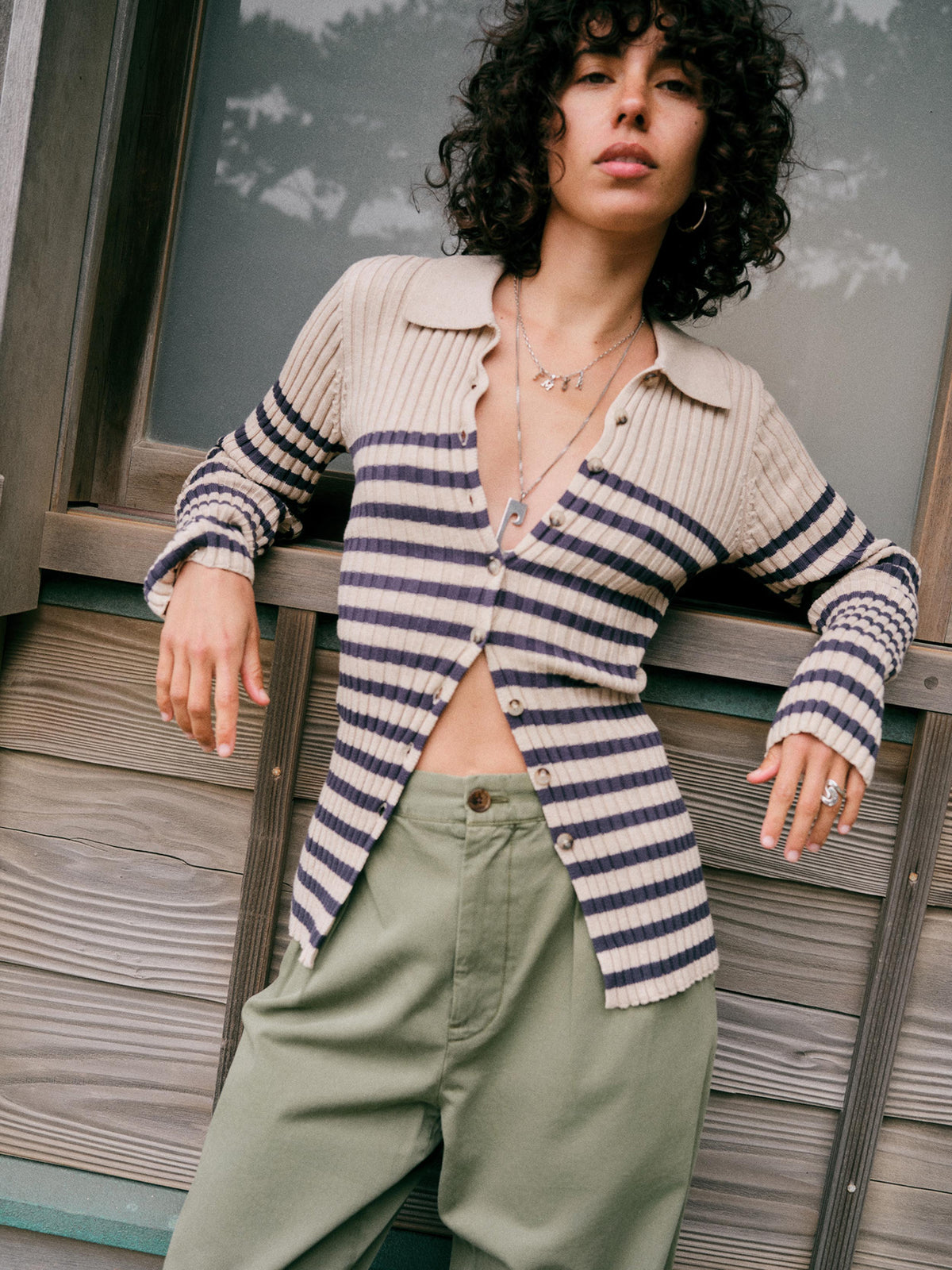 A model with curly black hair leans against a rustic wooden wall, wearing a beige and navy blue horizontally striped ribbed knit cardigan and sage green pants., image 1 of 7