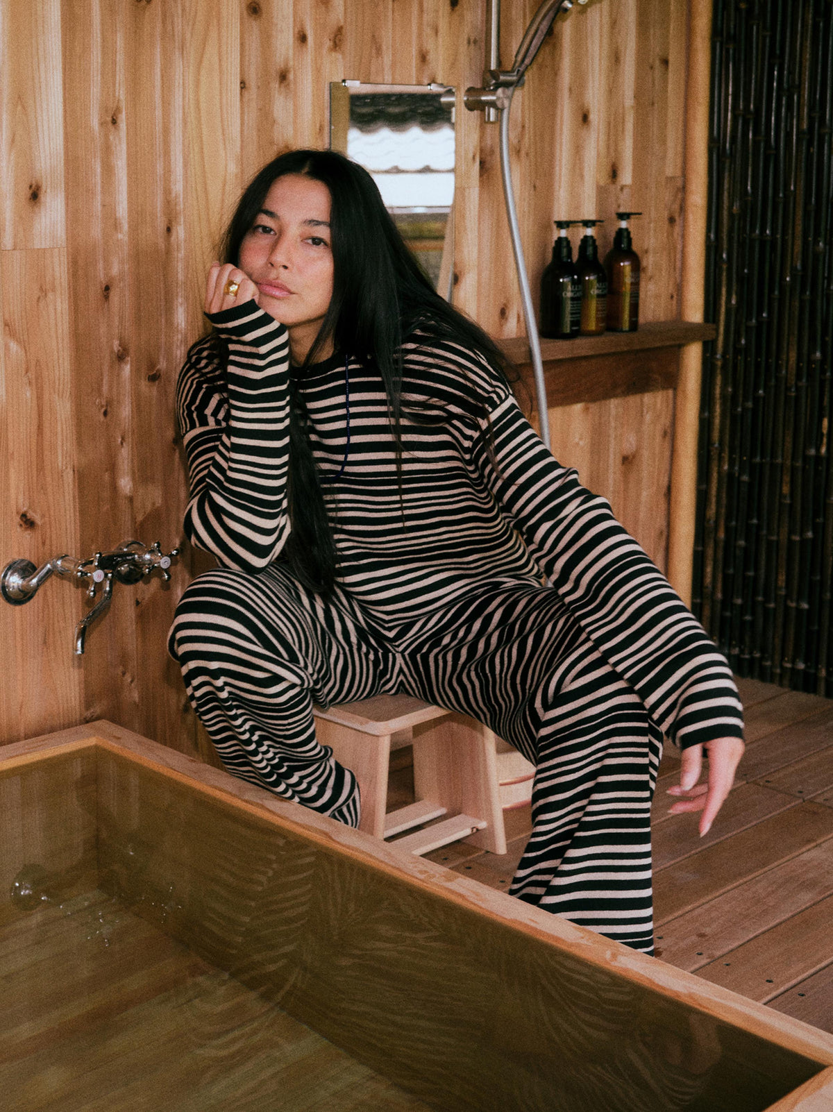 A woman with long black hair sits on a small wooden stool in a rustic wooden bathroom, wearing a matching black and beige striped lounge set. She is positioned next to a wooden bathtub, with a wooden wall, shower fixtures, and shampoo bottles in the background., image 1 of 6