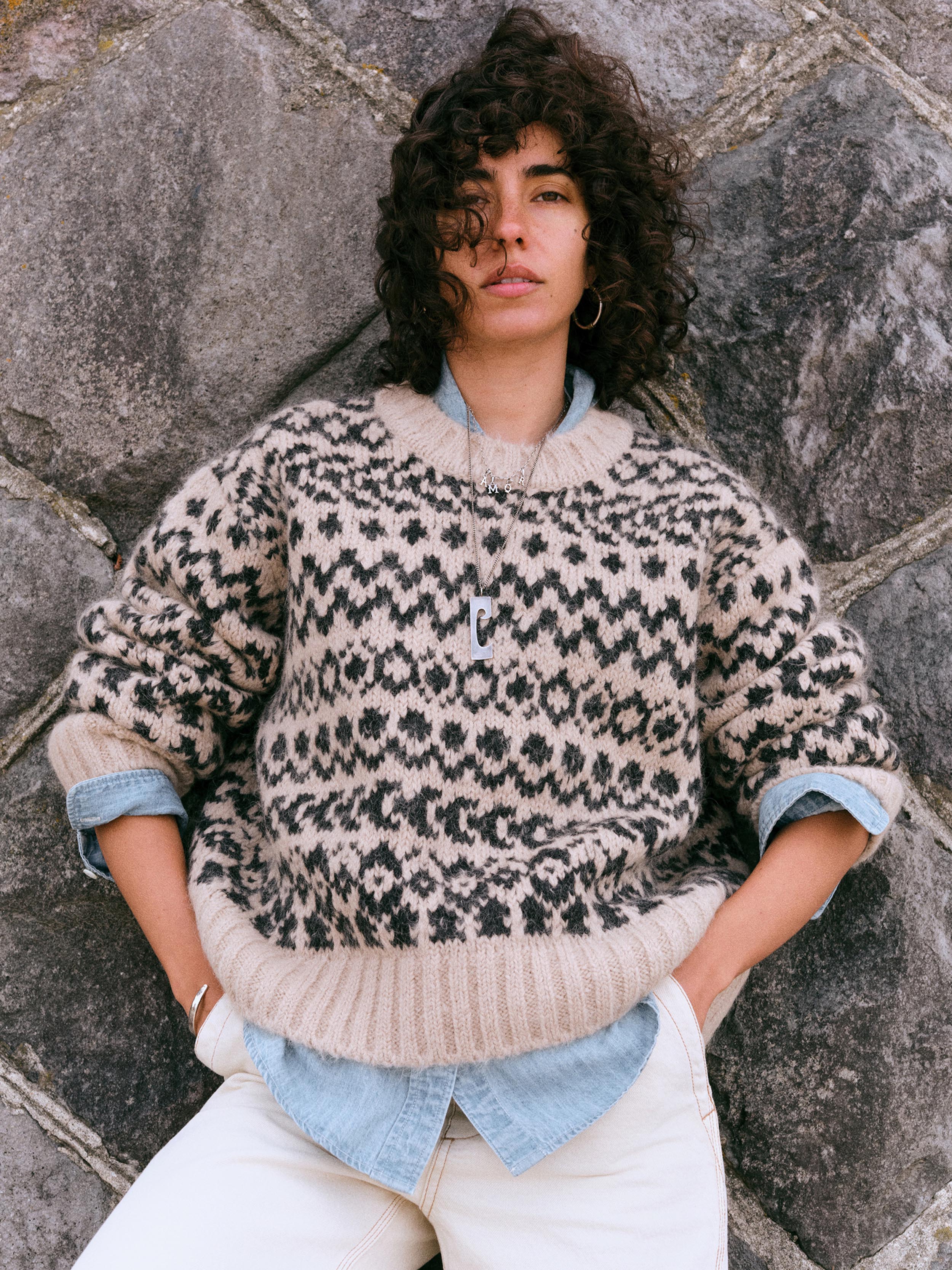 A model with curly dark hair wearing a beige and black patterned knit sweater over a light blue shirt, paired with white pants, leaning against a gray stone wall.