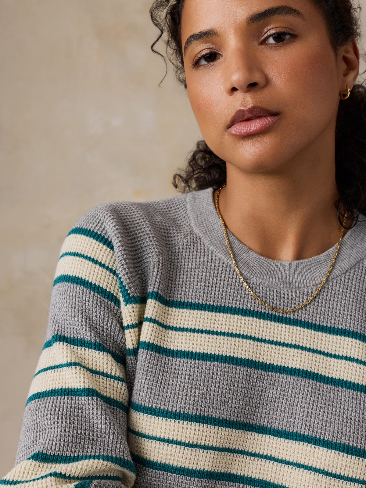 A close-up of a woman wearing a gray waffle-knit sweater with horizontal cream and teal stripes, paired with a delicate gold chain necklace., image 2 of 5