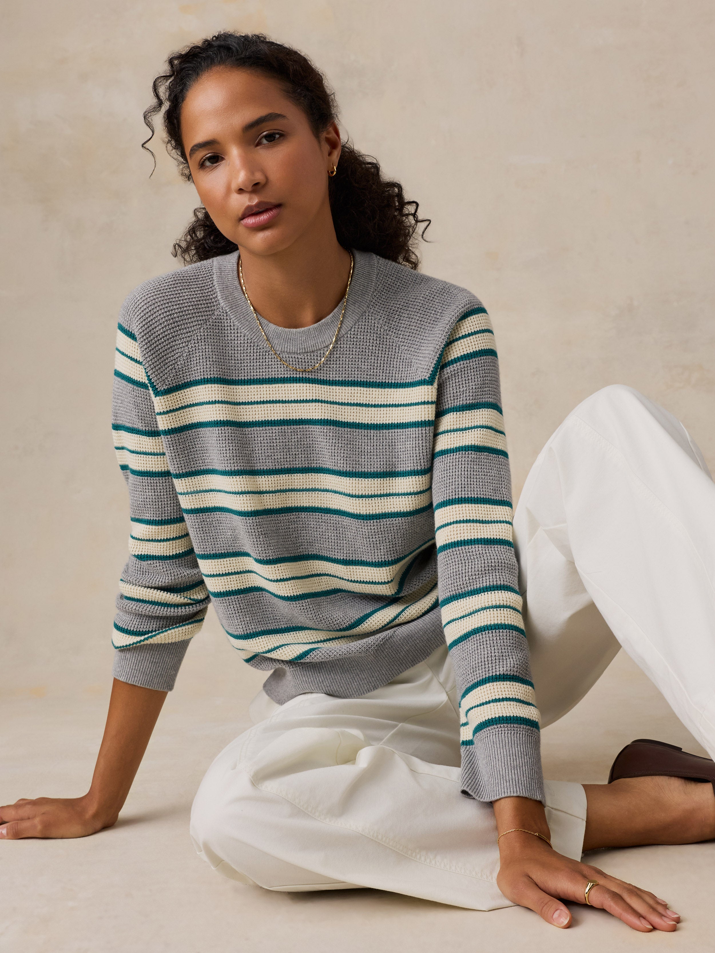 A woman sits on a beige floor against a textured beige wall, wearing a gray knit sweater with horizontal teal and cream stripes, paired with white wide-leg pants.