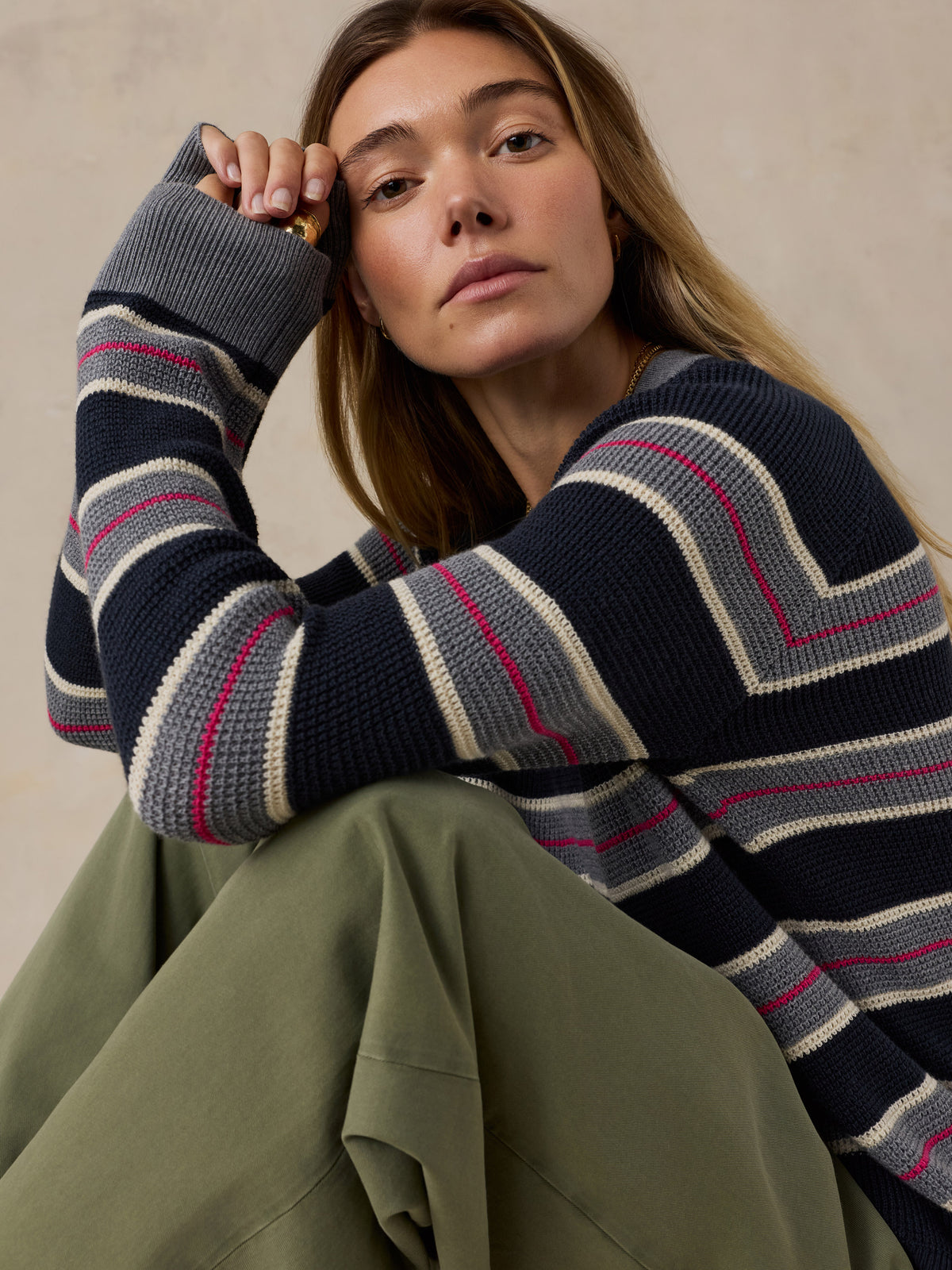 A woman wearing a navy blue knit sweater with horizontal stripes in beige, gray, and pink, paired with an olive green skirt, posed against a neutral beige background., image 2 of 5