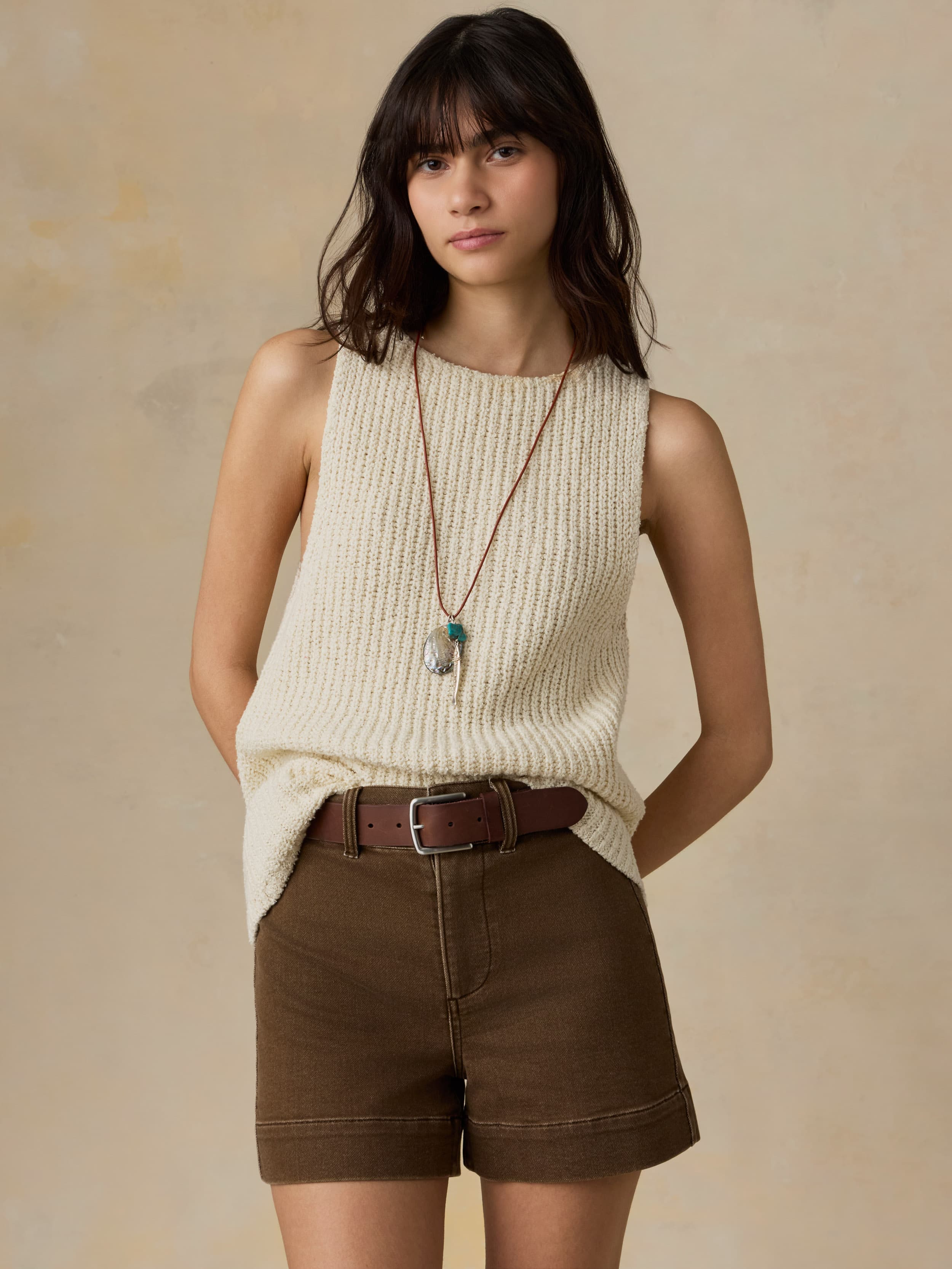 Woman wearing a cream sleeveless top and brown shorts against a beige background