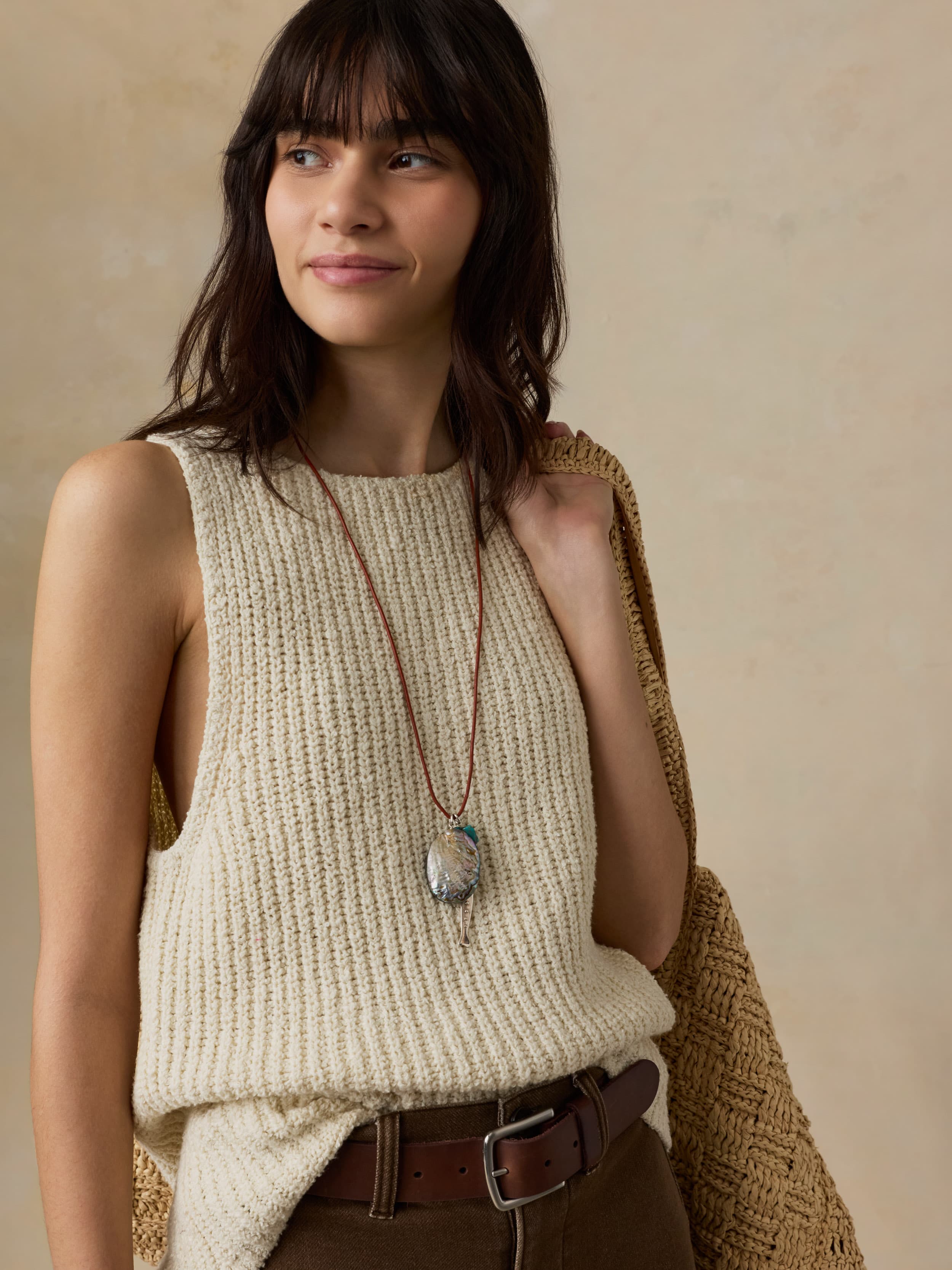 Woman wearing a beige knitted sleeveless top with a necklace, standing against a neutral background.