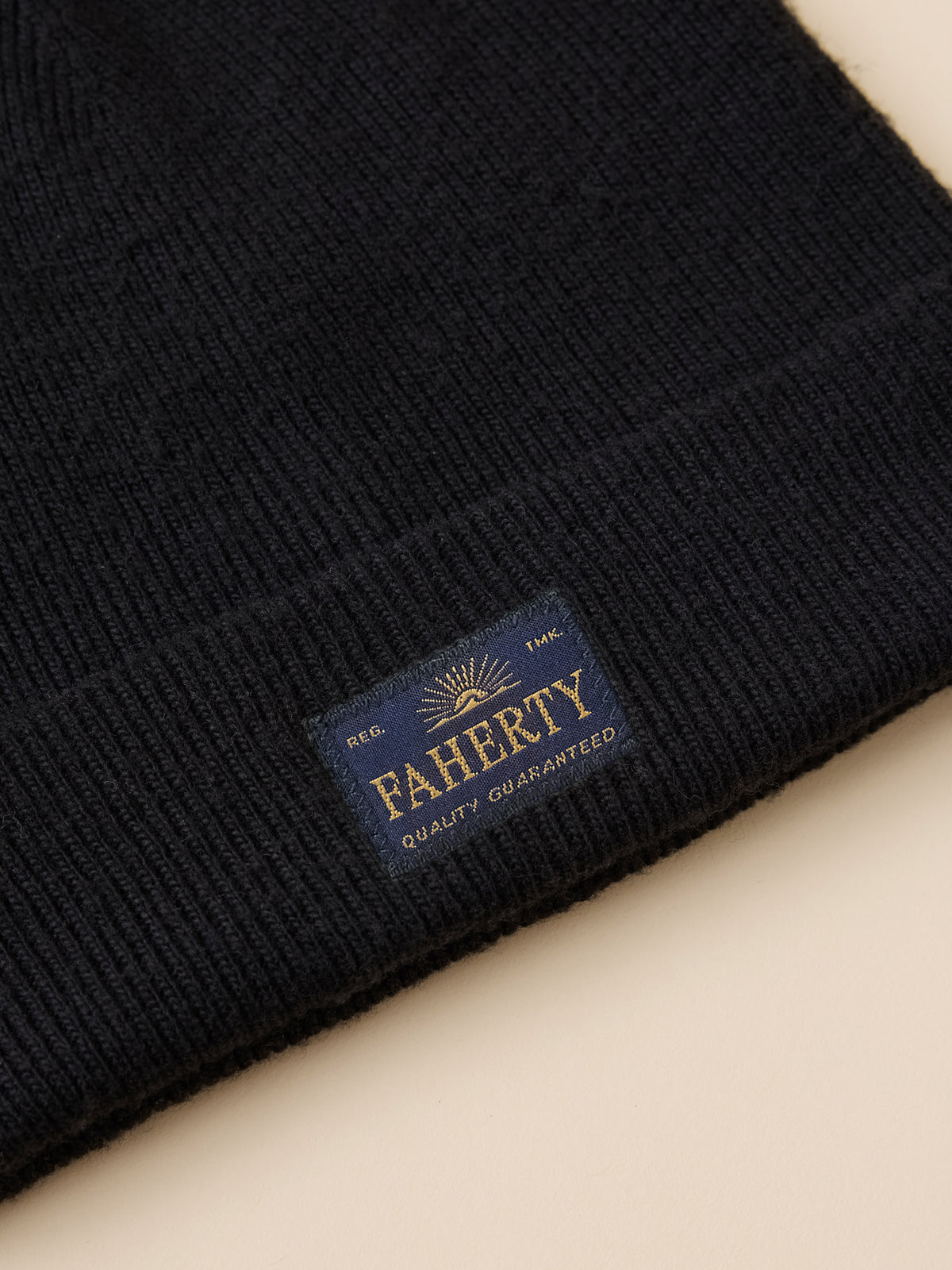 Close-up of a black ribbed knit beanie with a folded cuff showing a navy blue woven label that reads FAHERTY Quality Guaranteed., image 2 of 3
