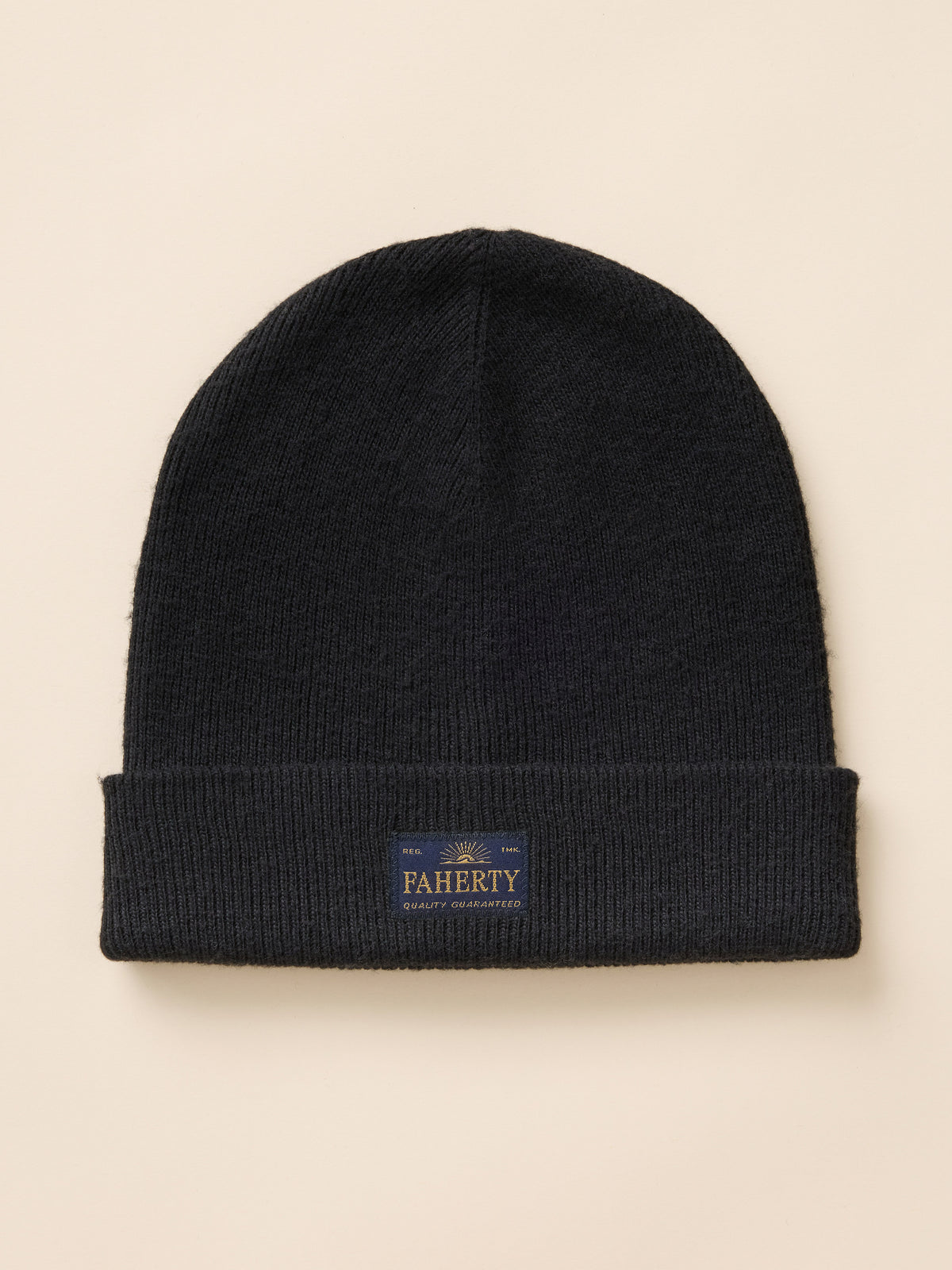 A black ribbed knit beanie hat with a folded cuff and logo patch, photographed flat against a light beige background., image 1 of 3