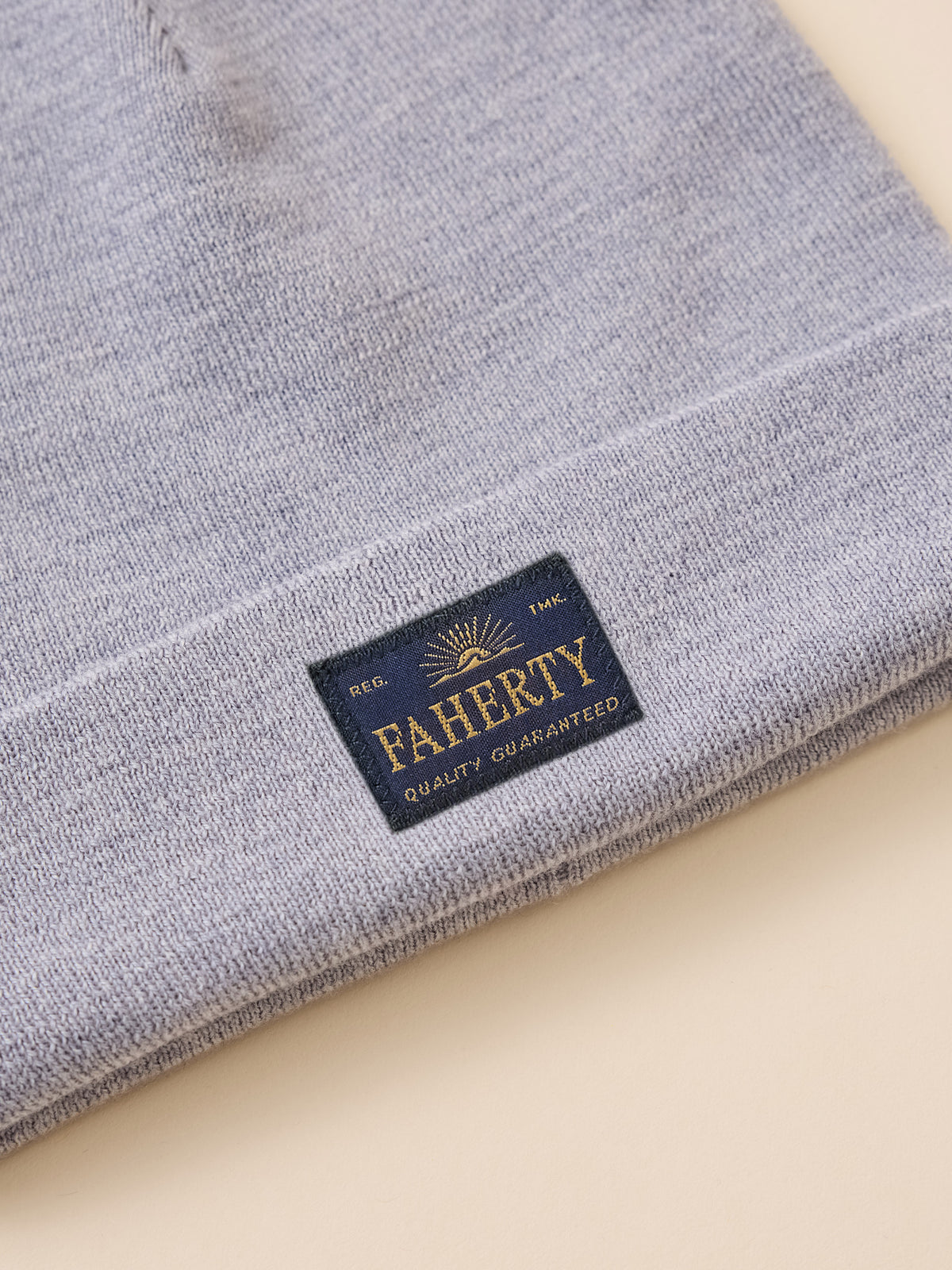 A close-up of a folded light heather gray knit item, likely a beanie or hat, showing a navy blue sewn-in label with gold lettering that reads FAHERTY and Quality Guaranteed., image 2 of 3