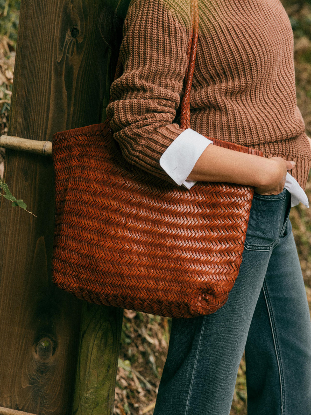 A woman wearing a brown ribbed knit sweater and blue jeans carries a large woven brown tote bag with a shoulder strap, standing beside a wooden post outdoors., image 2 of 4
