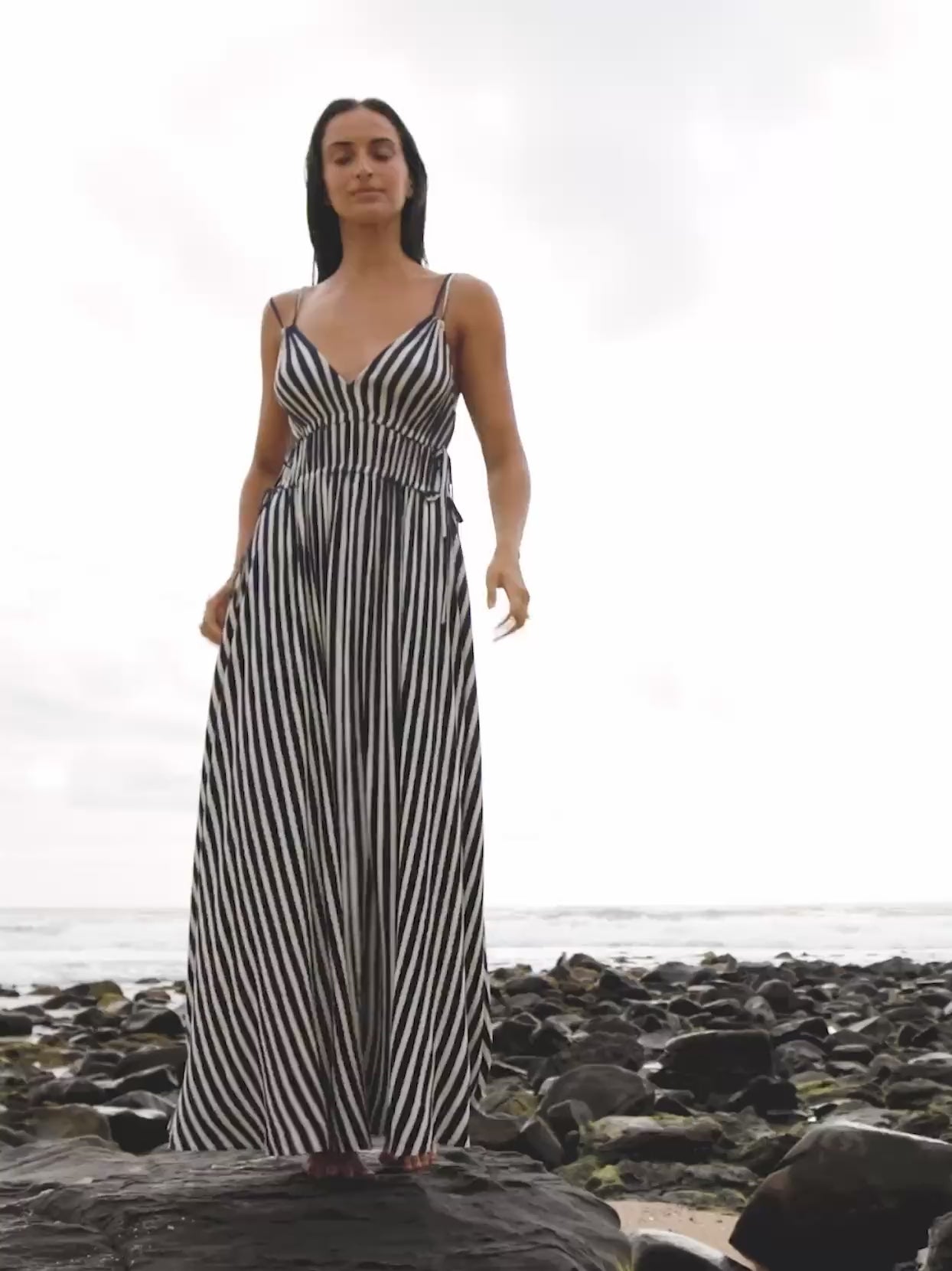 Video of a woman wearing a blue and white striped dress standing on rocks at the beach