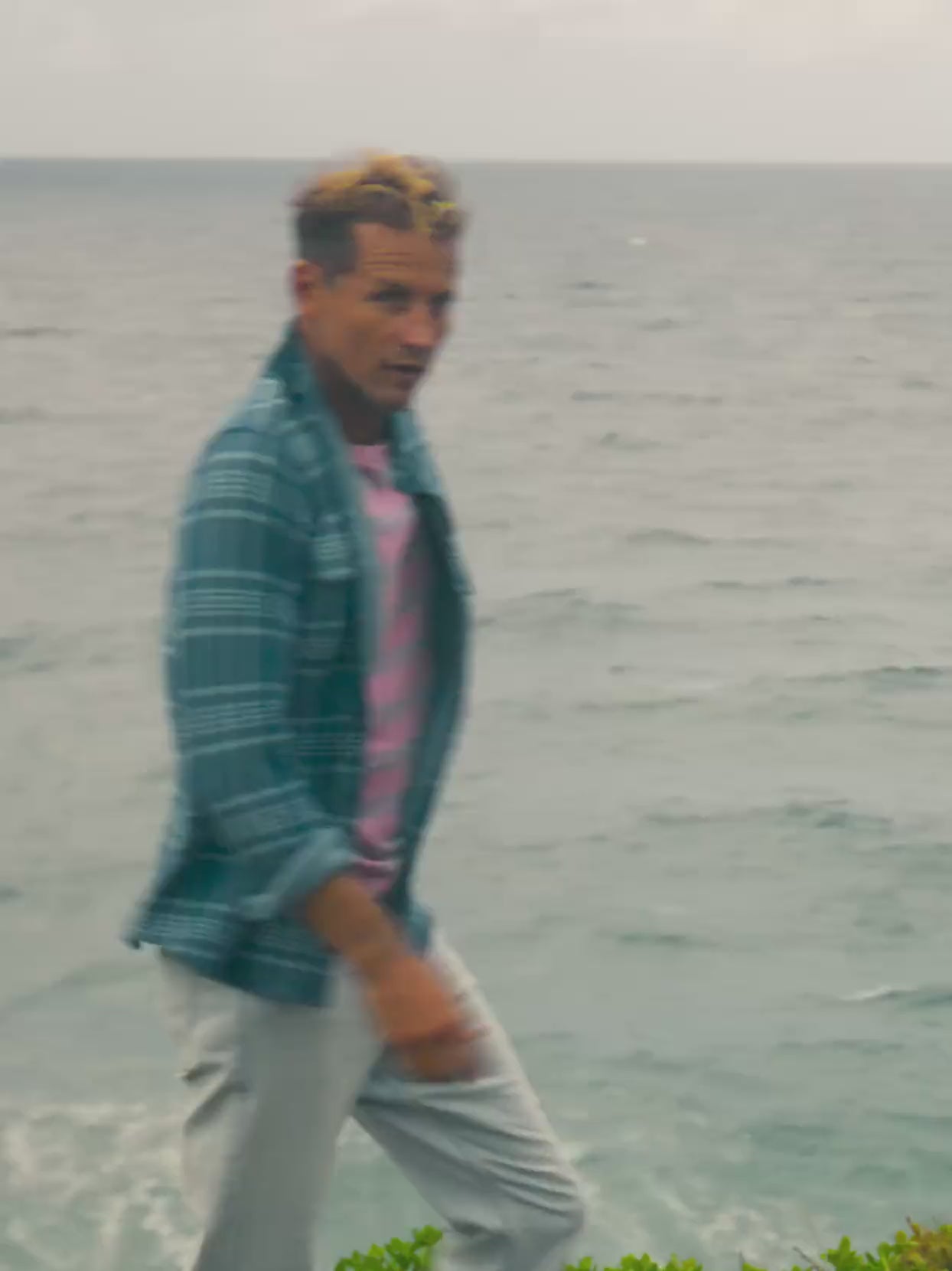 Video of a man wearing a pink t-shirt with a blue plaid shirt on top near the ocean