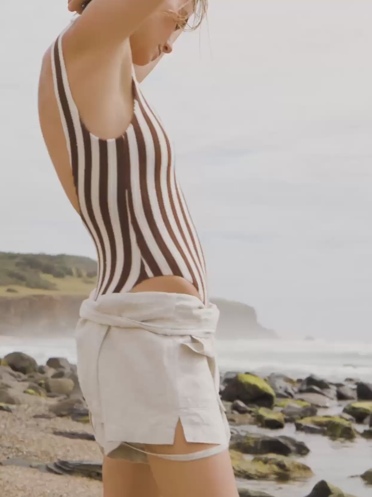 Video of a woman wearing a linen romper over a brown and white striped one piece swim suit standing on the beach