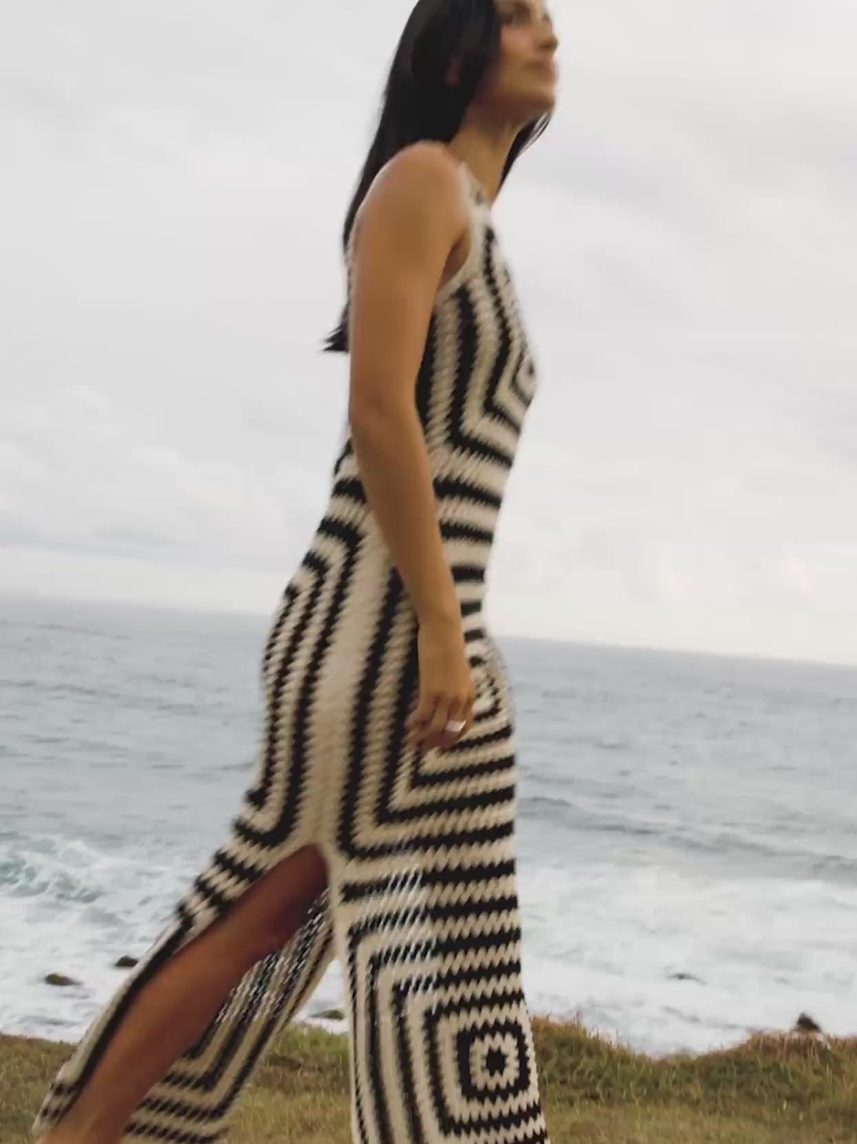 Video of a woman wearing a black and white crochet dress walking near the ocean