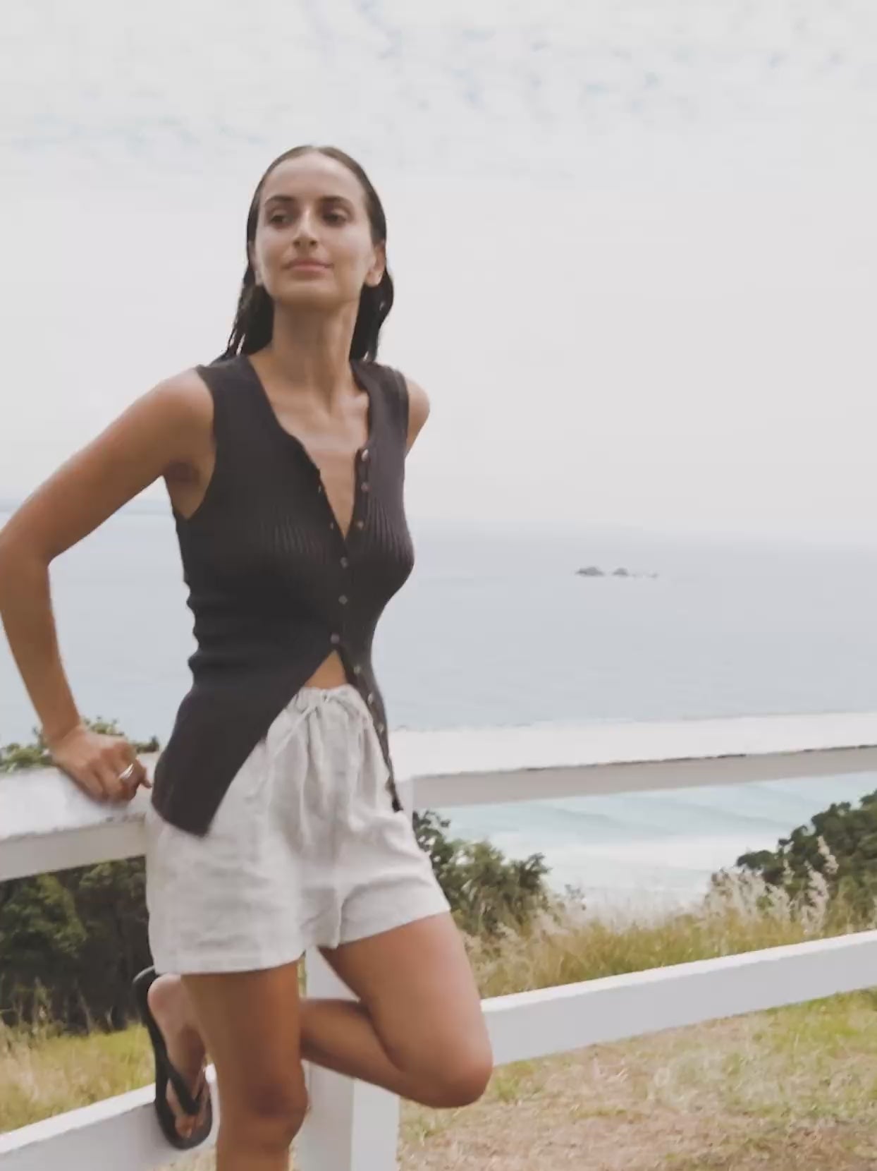 Video of a woman wearing a brown tank top with flax linen shorts sitting on a fence overlooking the ocean