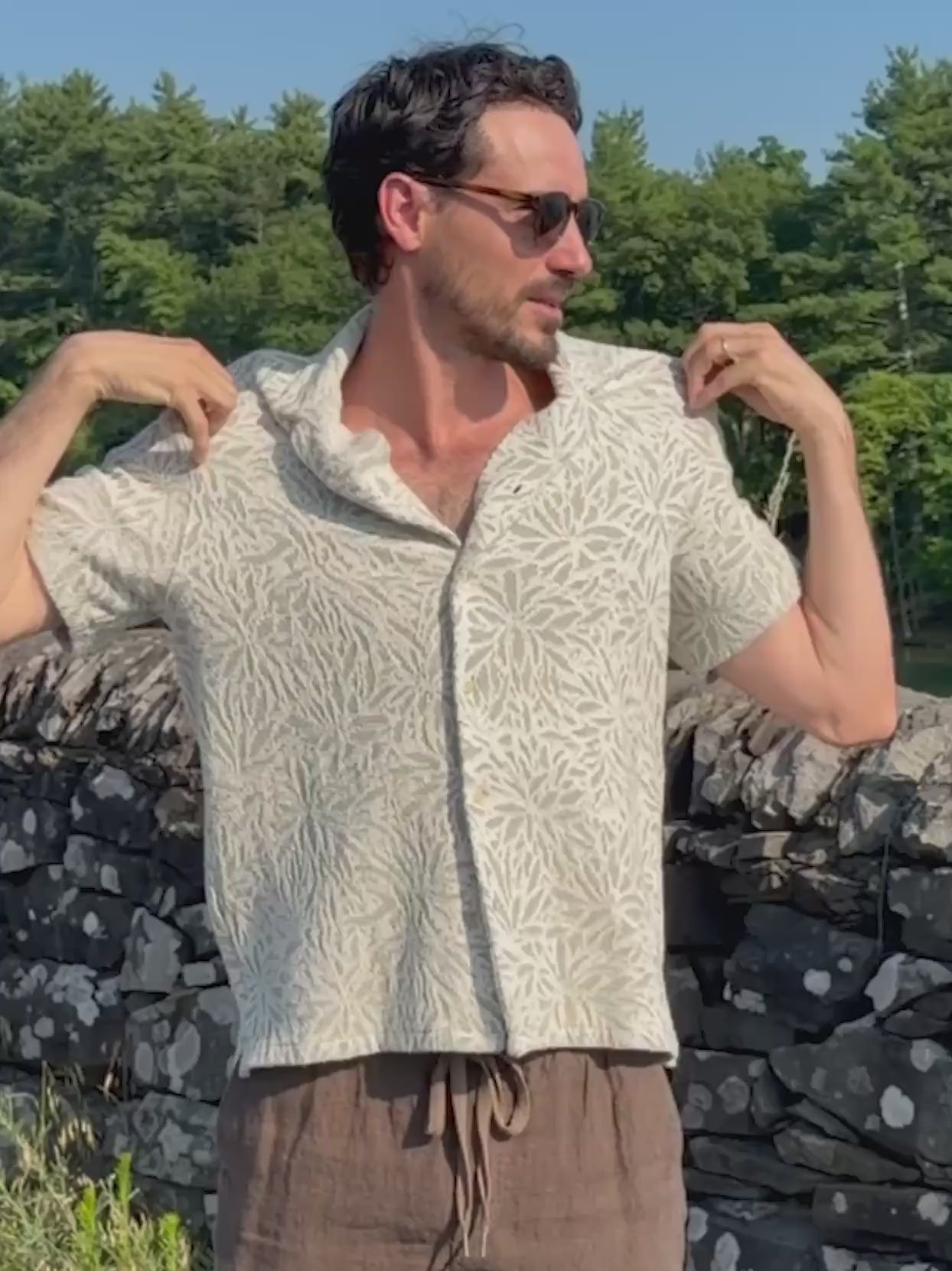 Man wearing a floral printed button up shirt and brown linen shorts standing outside in nature
