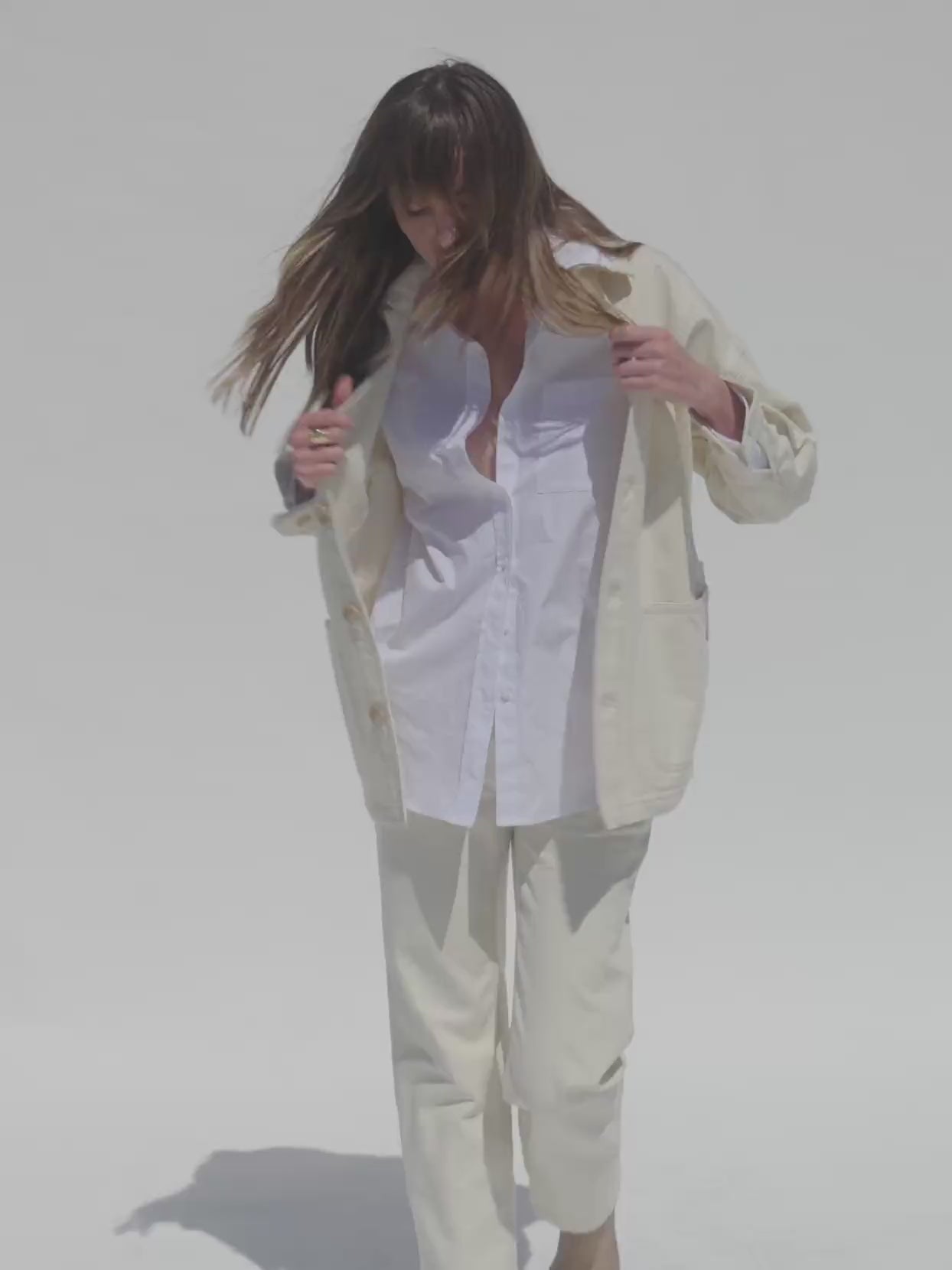 Video of a woman wearing a white button shirt, off white chore jacket, and matching pants against a white background