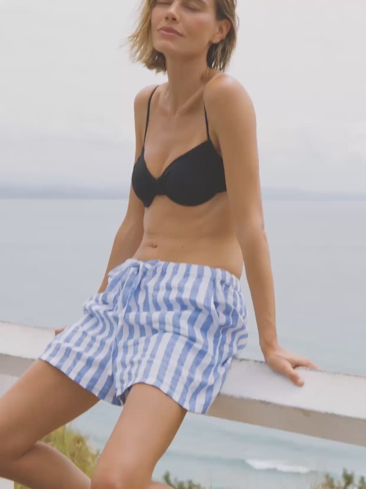 Video of a woman wearing a black swim suit top with blue and white striped linen shorts sitting on a fence overlooking the ocean