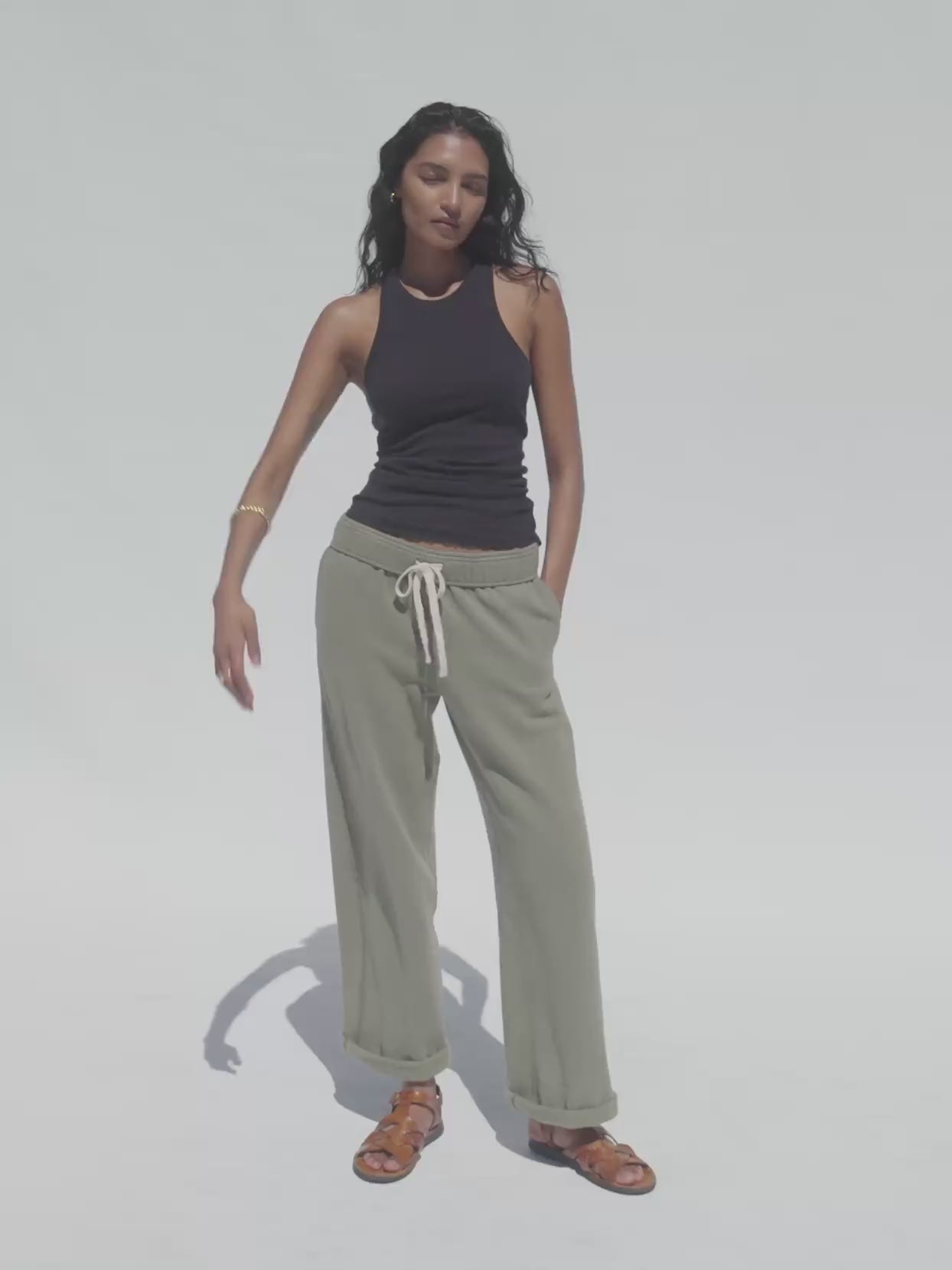 Video of a woman wearing a black tank top and olive green sweatpants against a white background