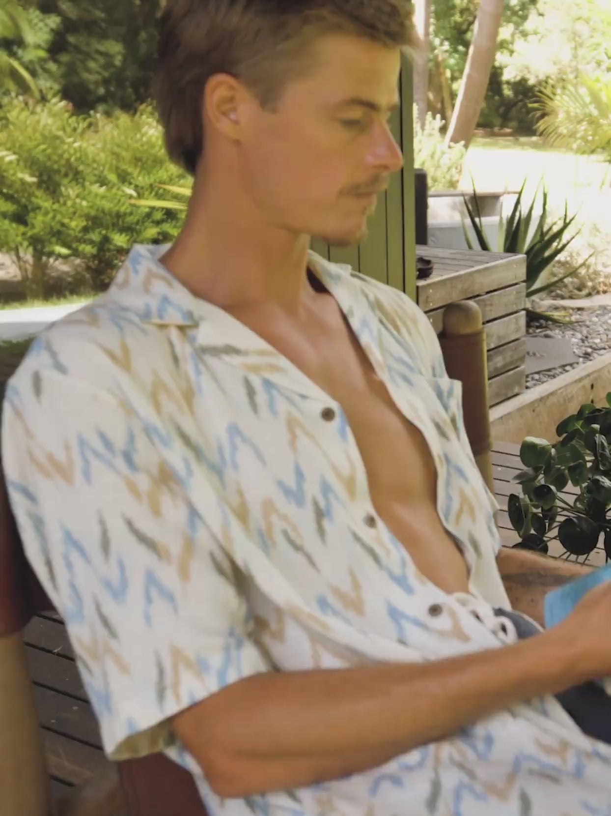 Video of a man wearing a printed short sleeve shirt sitting in a chair outside