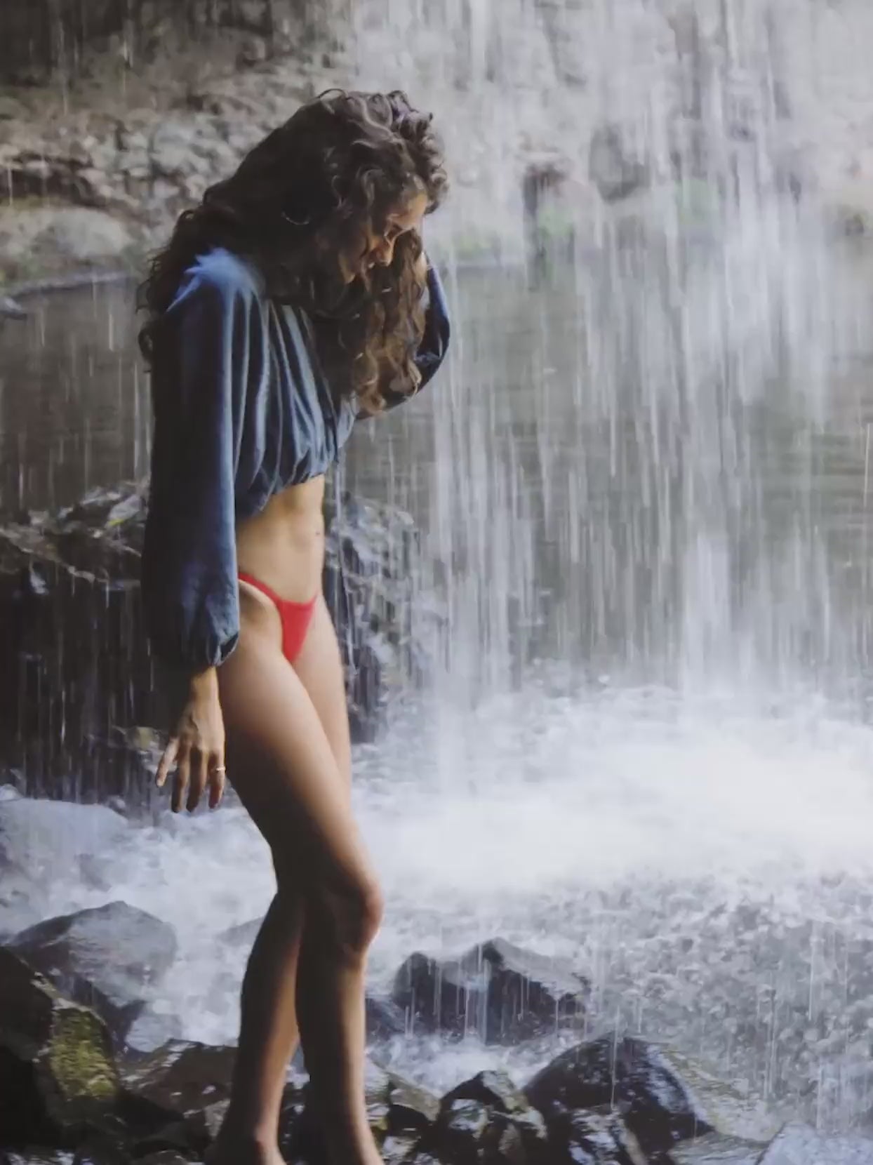 Video of a woman wearing a chambray top and red swim suit standing under a waterfall