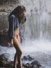 Video of a woman wearing a chambray top and red swim suit standing under a waterfall