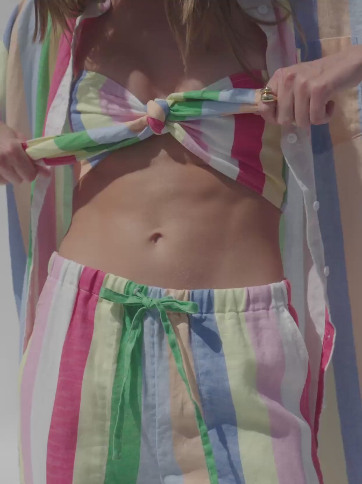 Video of a woman wearing multicolor striped bandeau top, matching pants, and a shirt dress against a white background