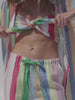 Video of a woman wearing multicolor striped bandeau top, matching pants, and a shirt dress against a white background