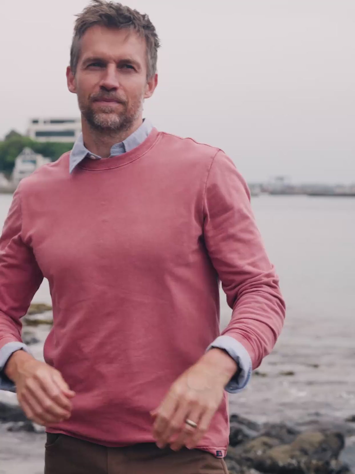 Video of a man wearing a red shirt over a blue button down shirt standing near the sea