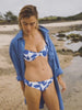 Video of a woman wearing a blue floral patterned swim suit and a blue linen shirt dress walking along the ocean