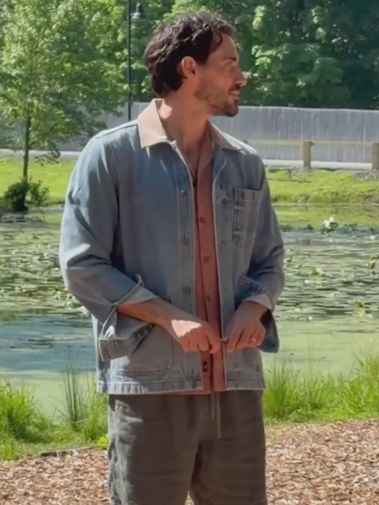 A man wearing a denim jacket over a button down shirt and brown linen shorts standing outside in nature