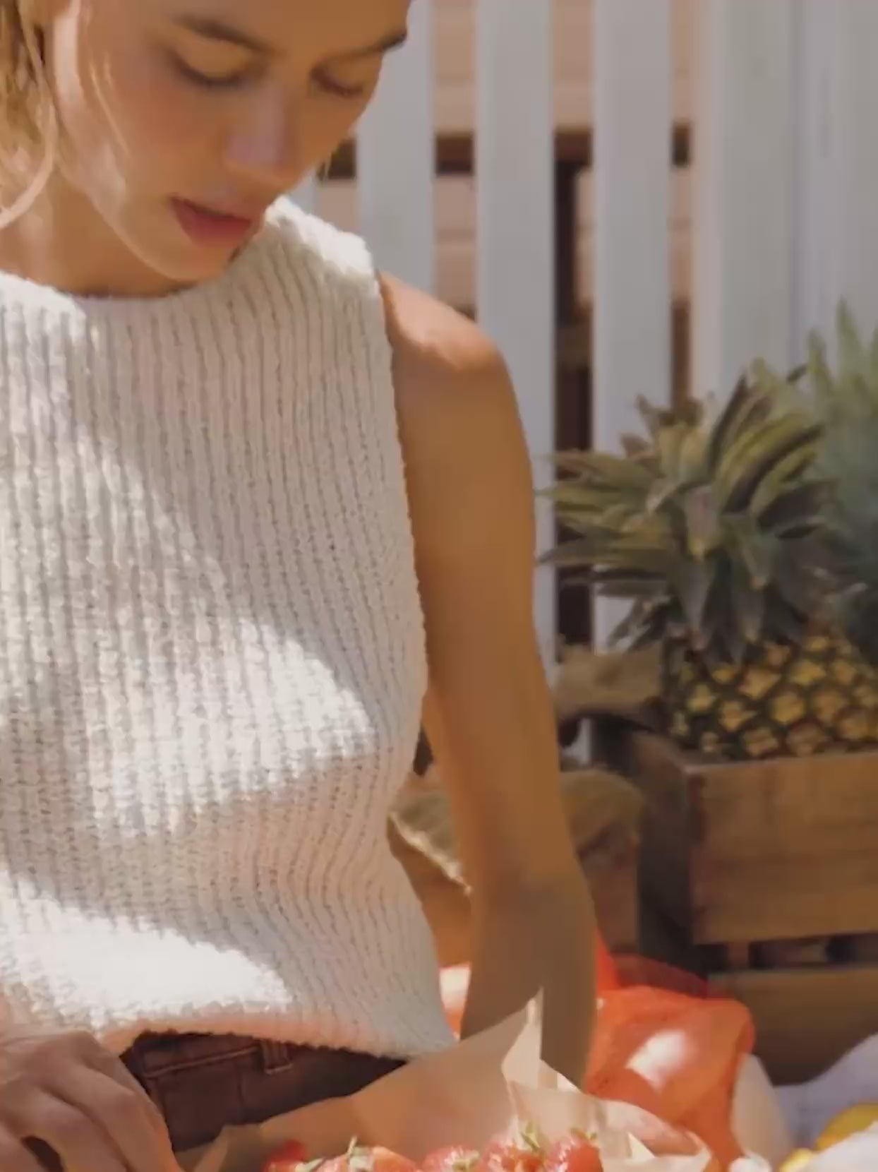 Video of a woman wearing a beige knit tank top holding strawberries at a farmer's market